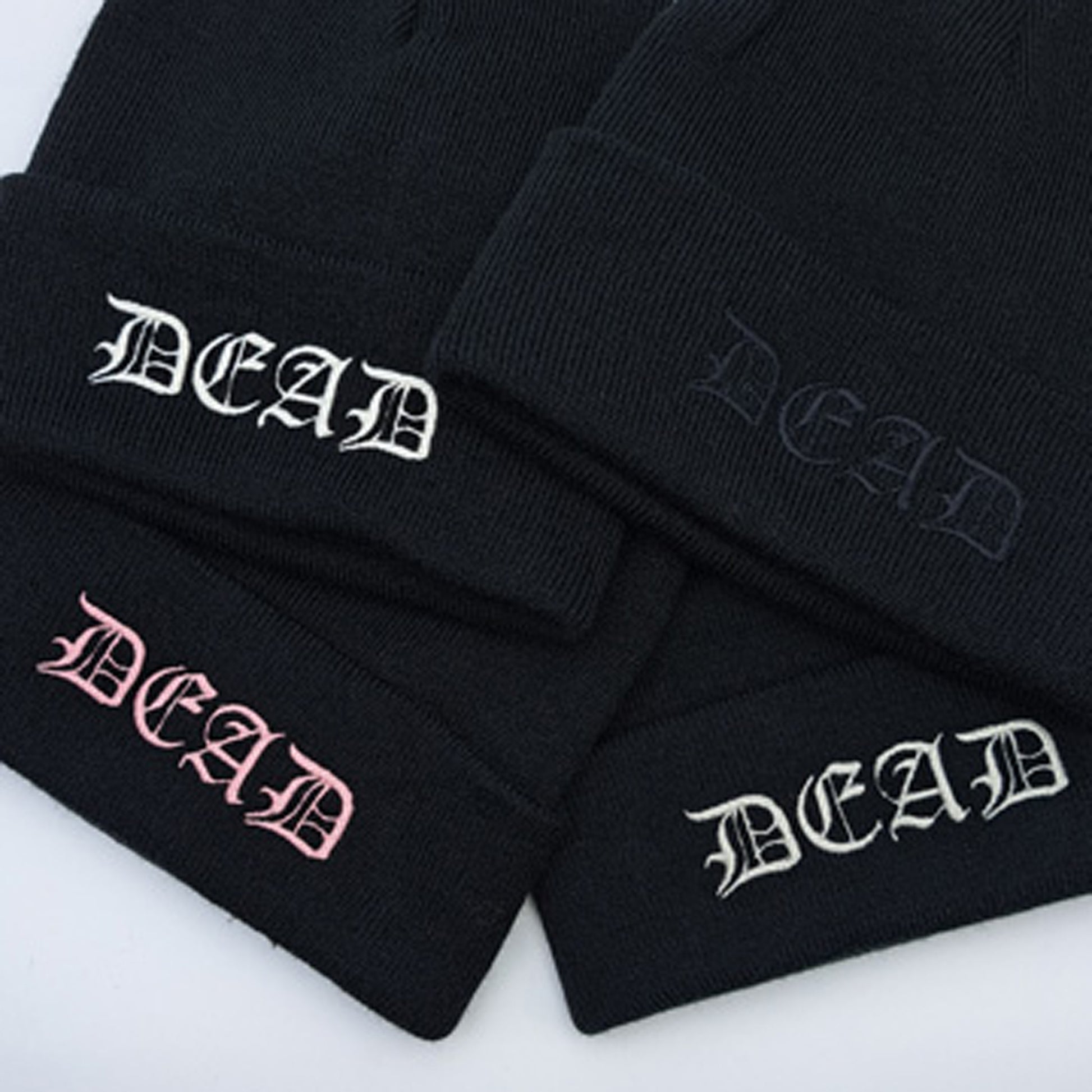 4 examples of BLACK knitted beanie hat featuring an embroidered cuff that reads DEAD in a medieval gothic font. Embroidery colours shown, white, black, pink and silver. Closer viewpoint.