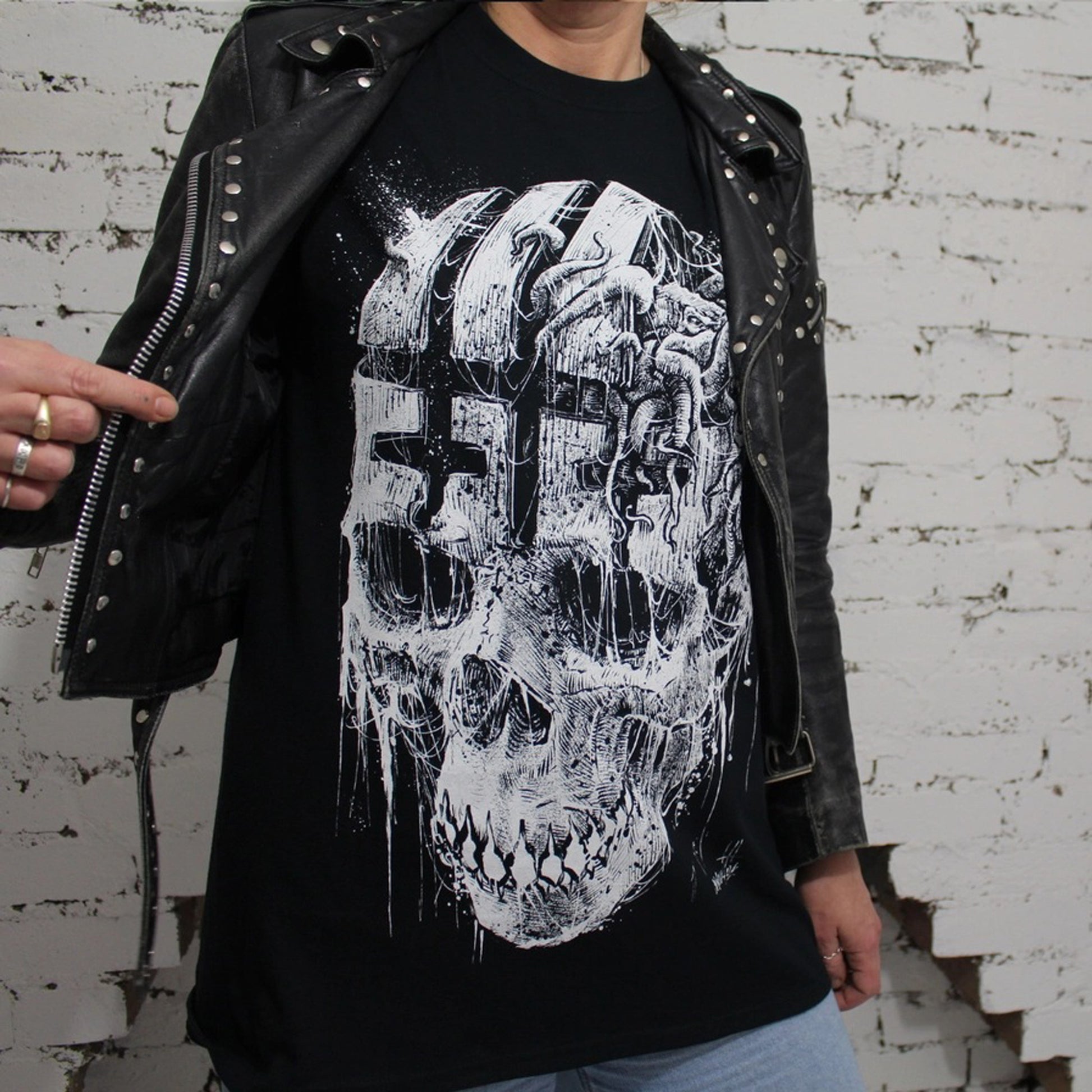 Lady in studded black leather jacket wearing this black t-shirt with large print of skull with a cryptid creature with tentacles and wing pushing out of the forehead region.