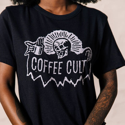 A person wearing a black short-sleeve graphic T-shirt with a white distressed skull, fanburst, coffee cup, and the words 'COFFEE CULT' on the front.