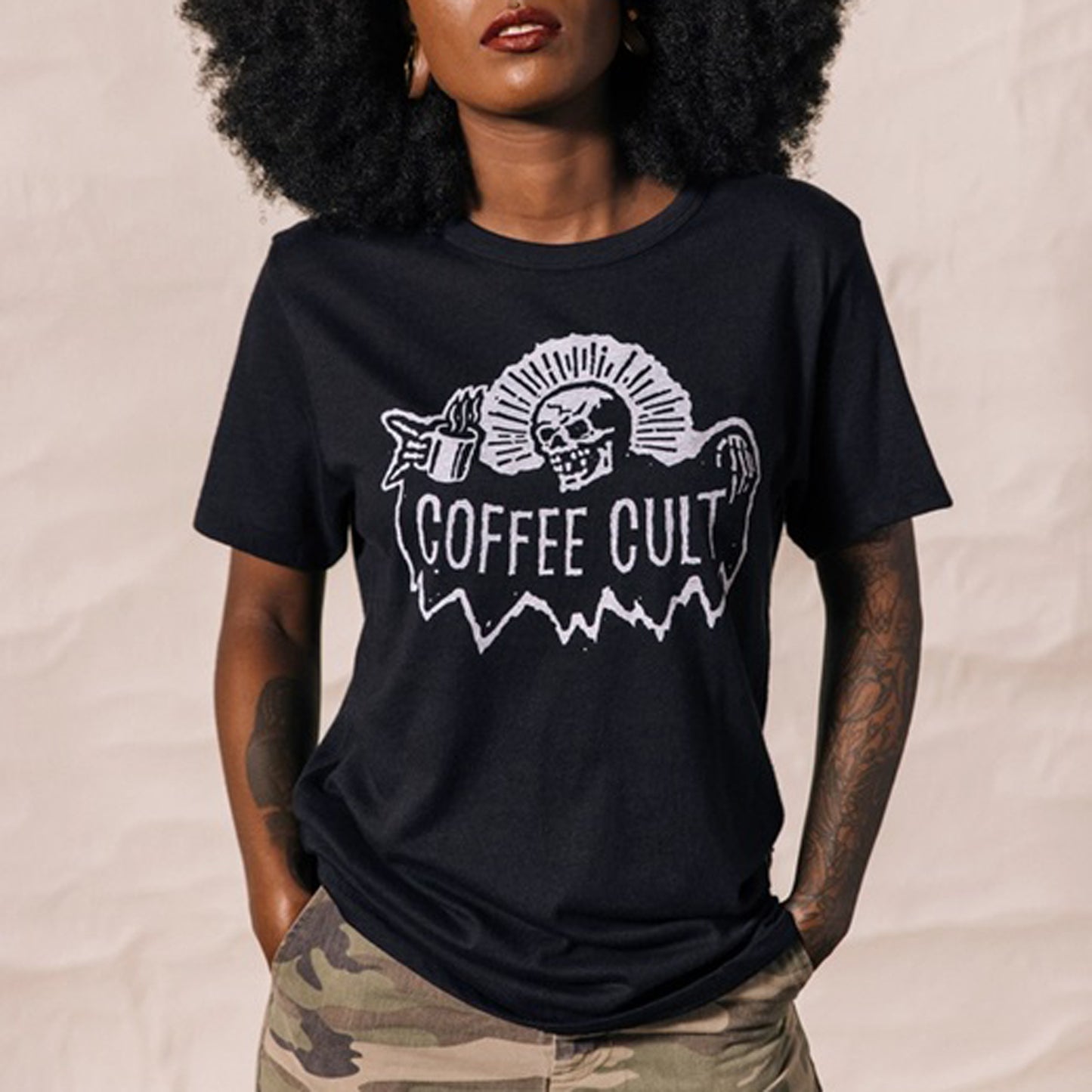 A woman wearing a black graphic t-shirt with a white 'COFFEE CULT' reaper skull design, paired with camouflage pants.