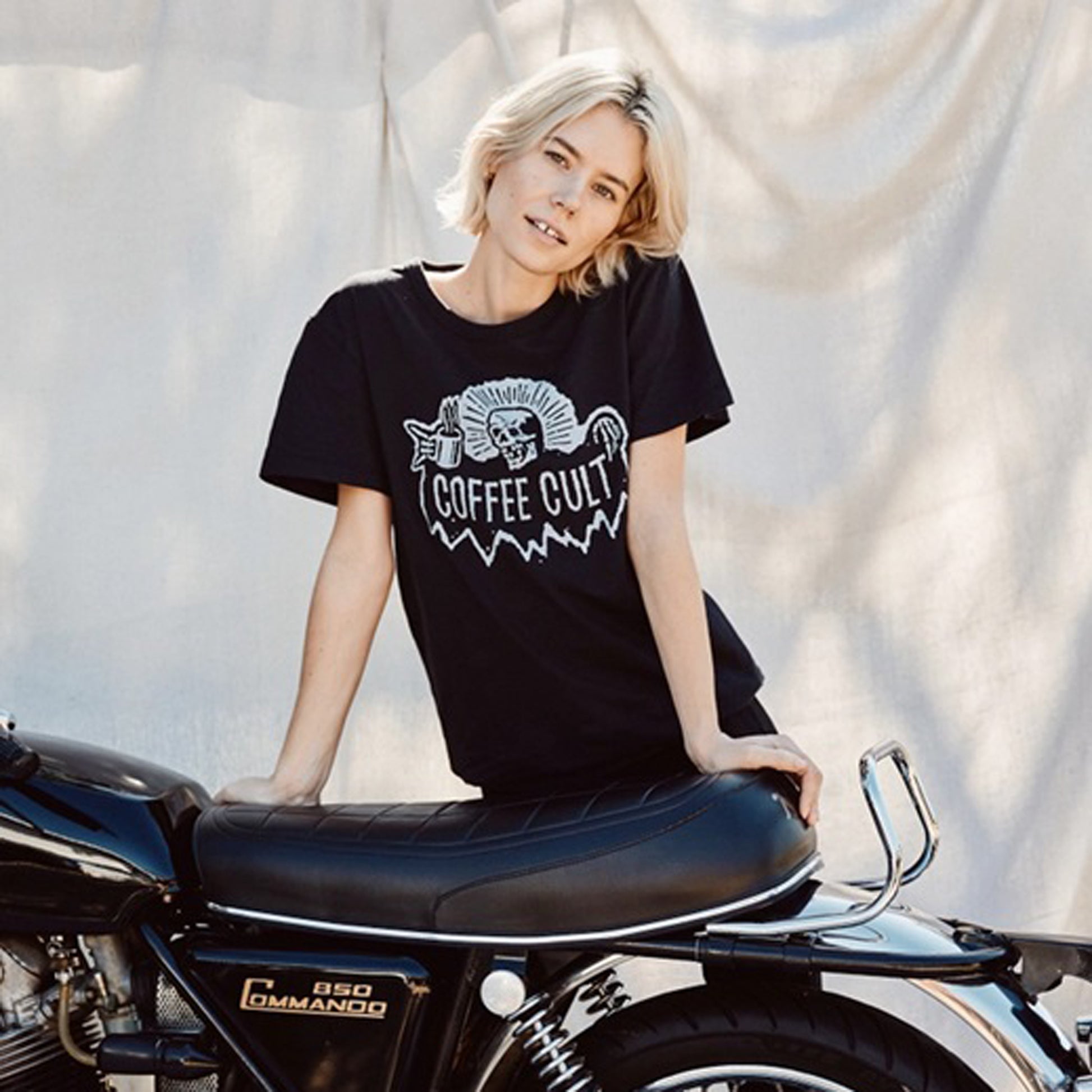 A woman with short blonde hair leans on a black motorcycle, wearing a black graphic t-shirt with a white 'Coffee Cult' reaper skull design.