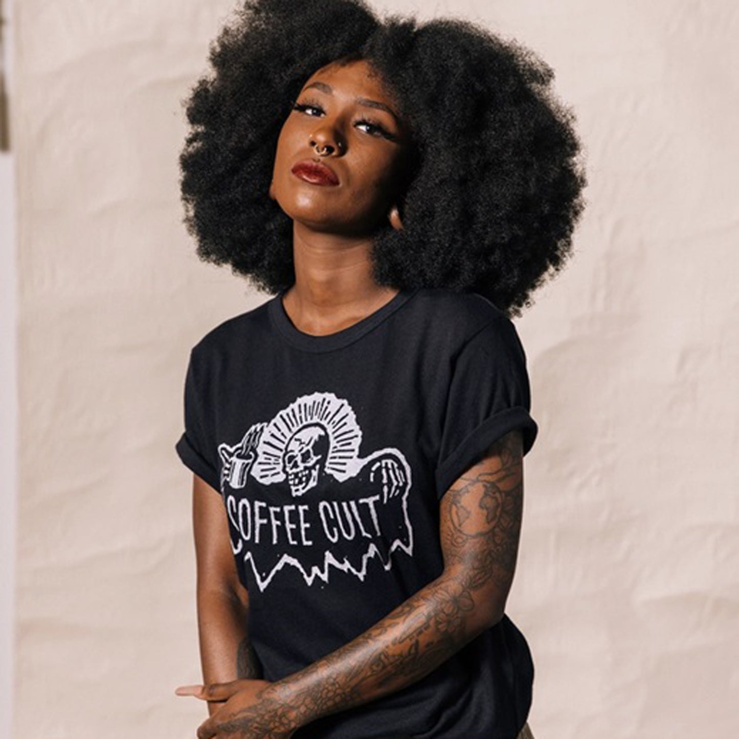 A woman with a large afro and tattoos wearing a black graphic t-shirt with a white 'COFFEE CULT' reaper skull design, posing against a wall.
