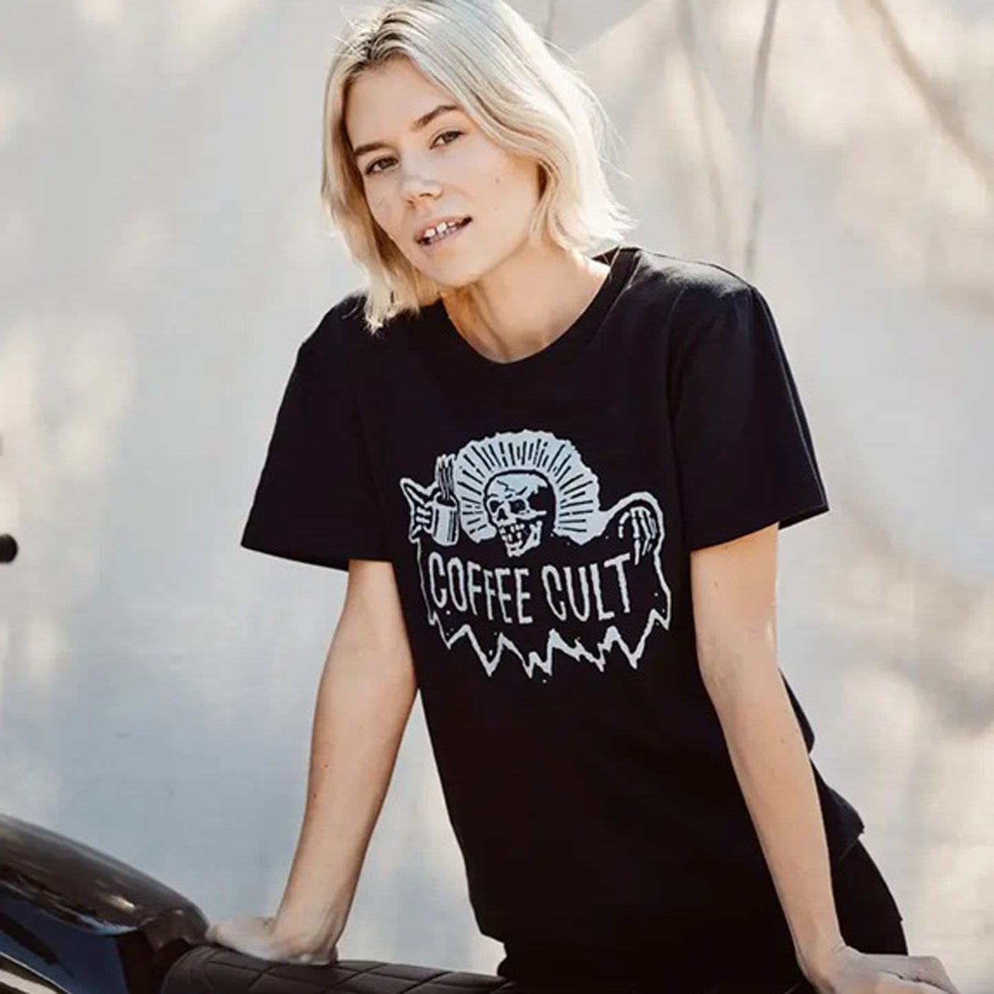 A woman with short blonde hair wearing a black graphic t-shirt with a white 'COFFEE CULT' reaper skull design, leaning against a motorcycle outdoors.