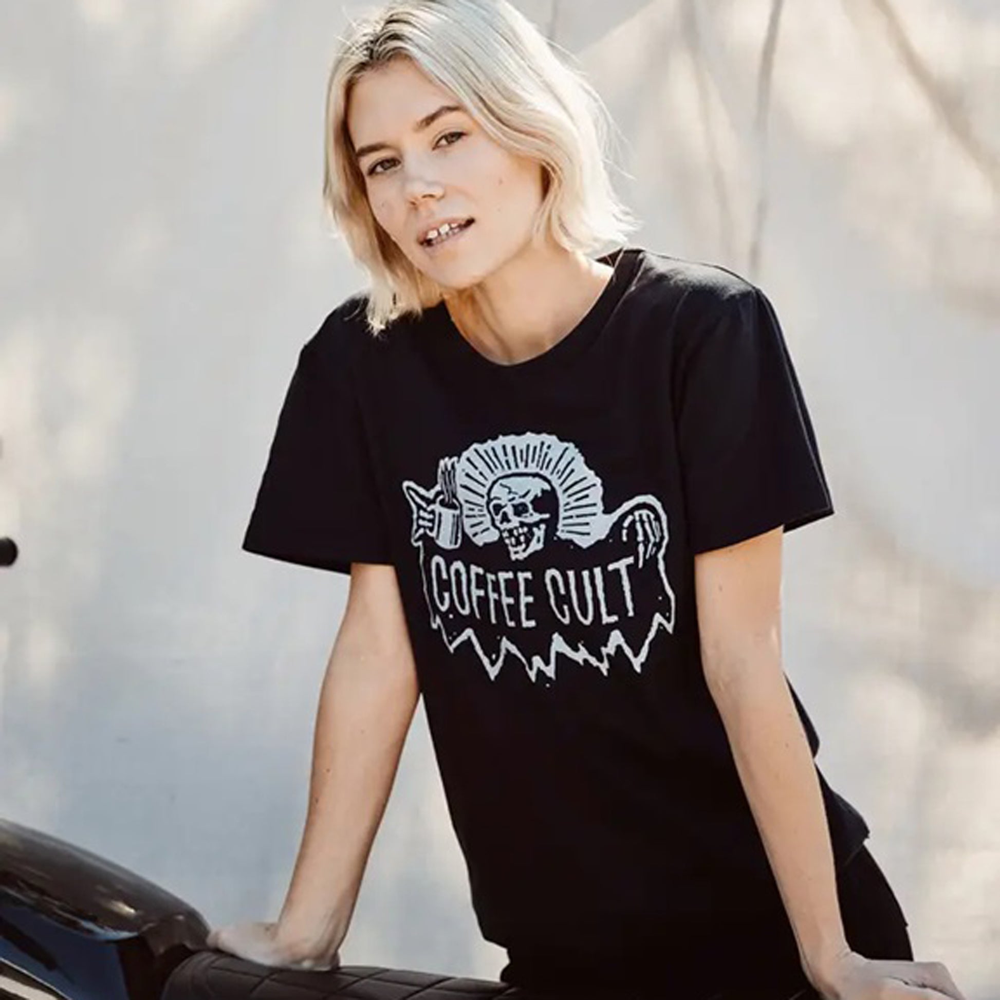 A woman with short blonde hair wearing a black graphic t-shirt with a white 'COFFEE CULT' reaper skull design, leaning against a motorcycle outdoors.