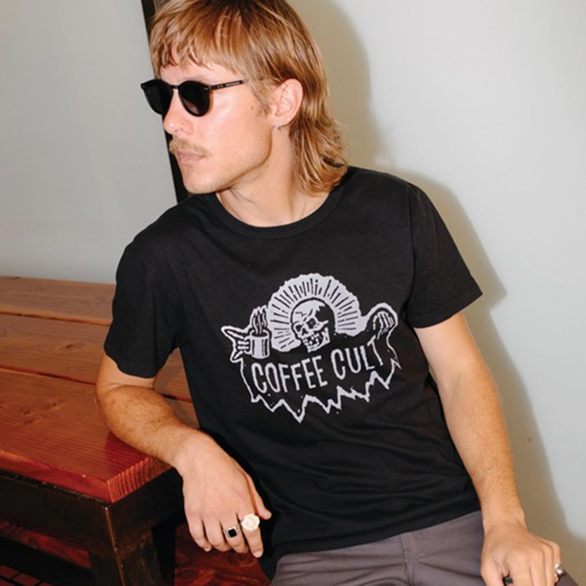 A man wearing a black graphic t-shirt with a white 'COFFEE CULT' reaper skull and mug design, sitting on a wooden bench against a plain wall.