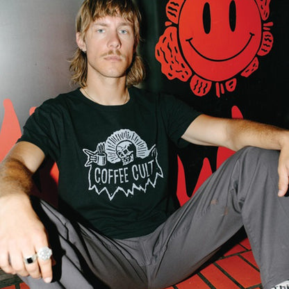 A man wearing a black graphic t-shirt with a white 'COFFEE CULT' skeleton reaper holding a mug design, sitting in front of a red smiley face wall.