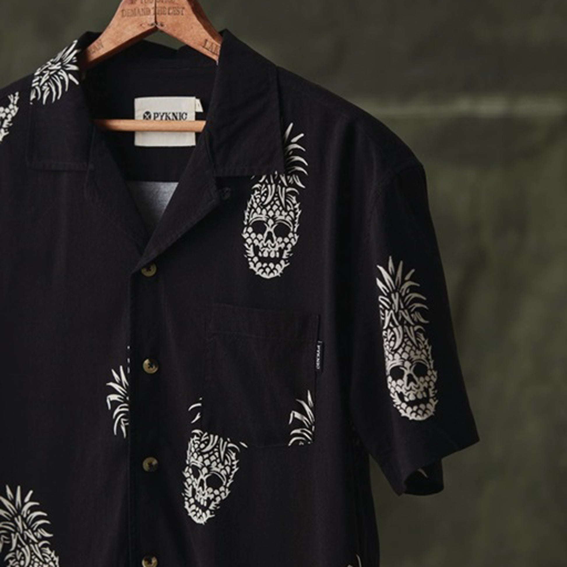 A black short-sleeve button-up shirt with white pineapple skull prints, hanging on a wooden hanger against a dark textured background. The brand name 'PYKNIC' is visible on the inner neck label.