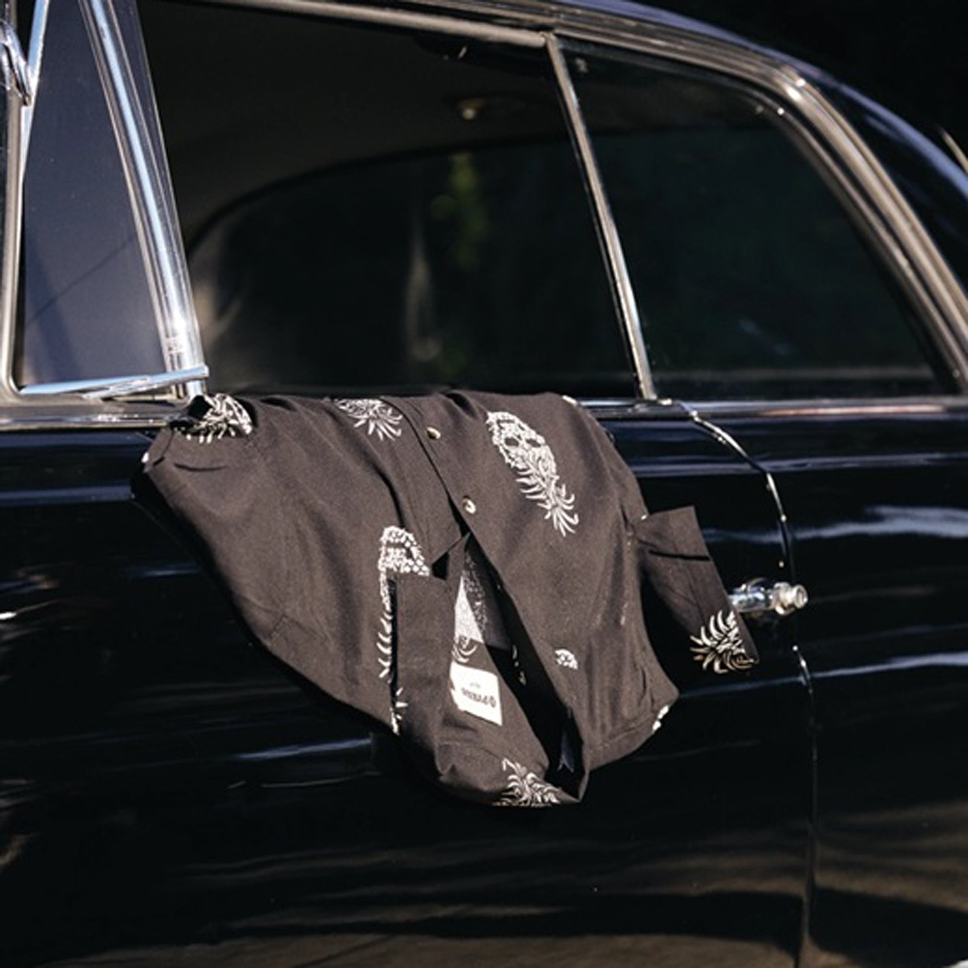 A black short-sleeve button-up shirt with white pineapple skull prints draped over the open window of a black vintage car.