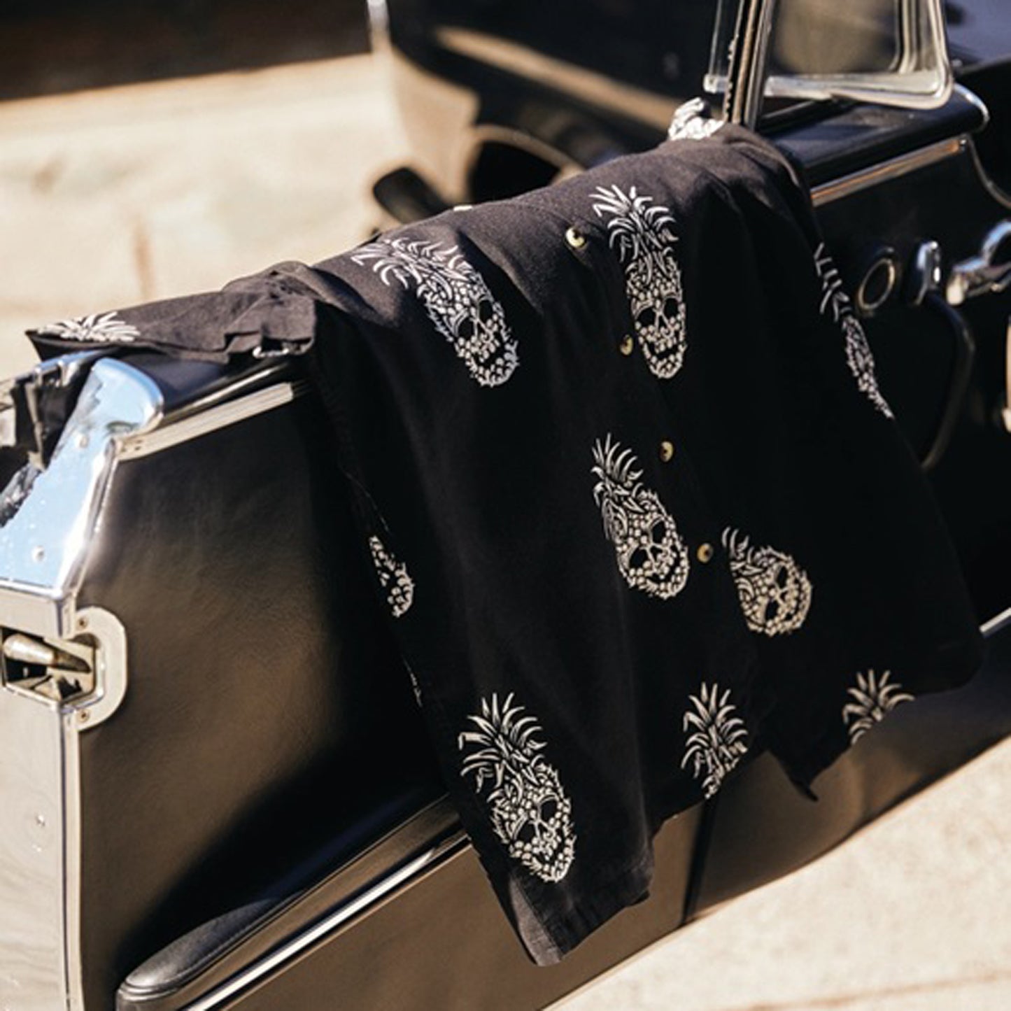 A black short-sleeve button-up shirt with white skull-and-pineapple prints, draped over the side of a vintage car outdoors.