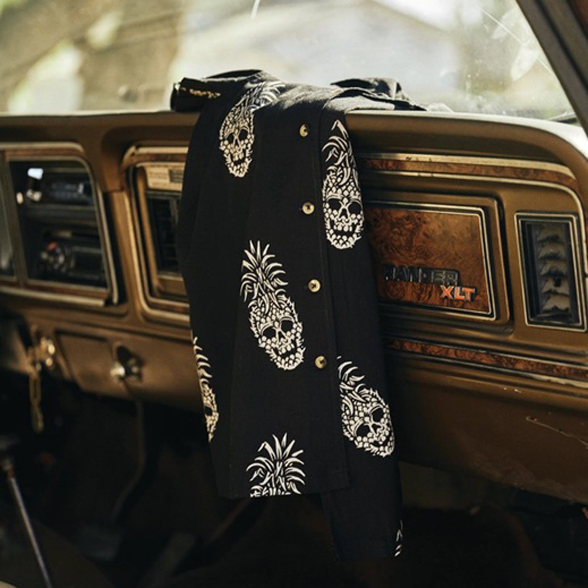 A black button-up shirt with white skull and pineapple prints draped over the dashboard of a vintage car interior.