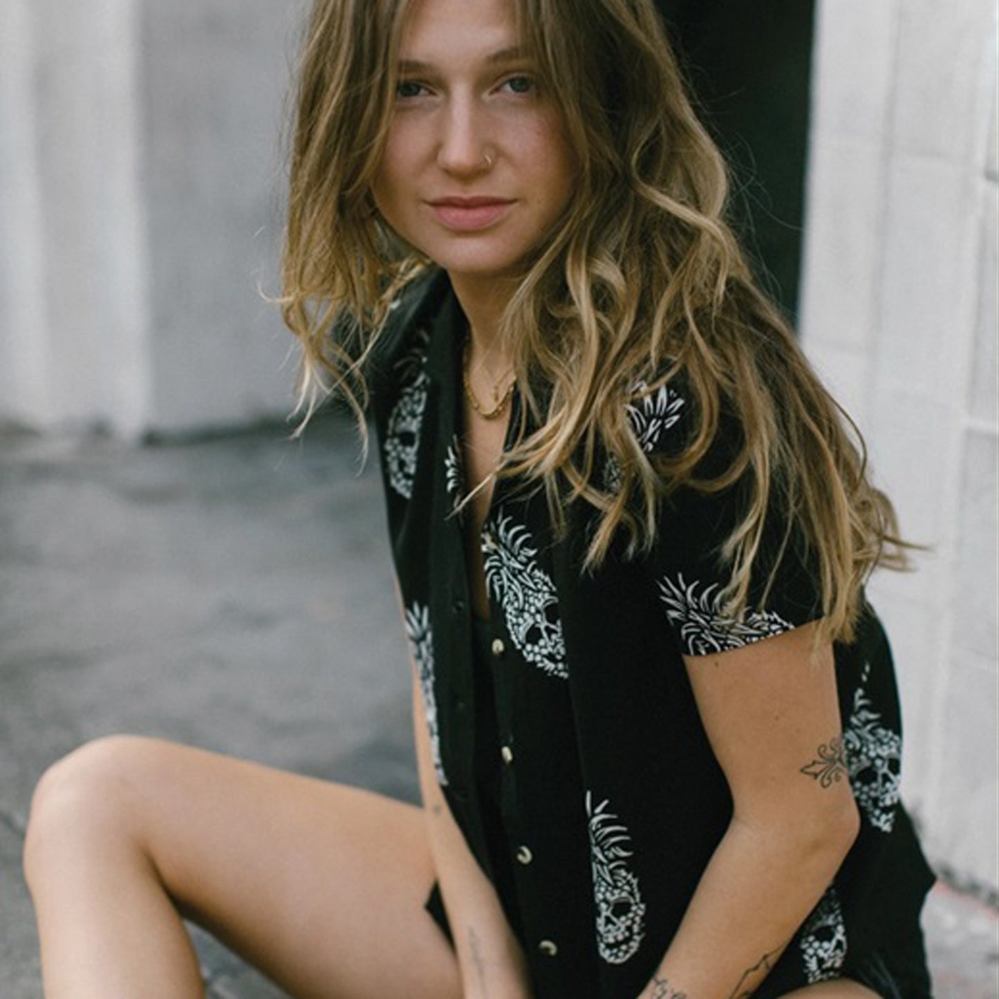 A woman with wavy light brown hair sits outdoors wearing a black short-sleeve button-up shirt with a white pineapple skull prints. She is looking directly at the camera against a blurred gray concrete background.