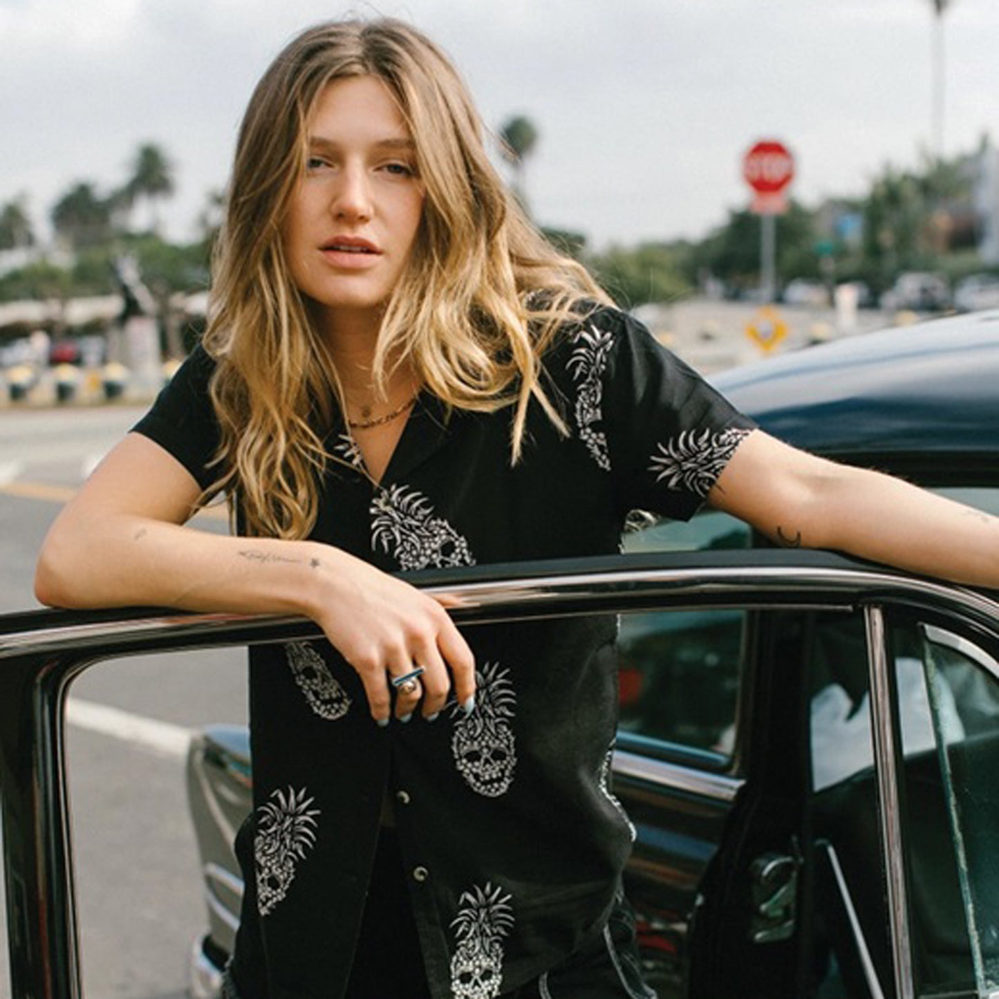 A woman leans on the open door of a vintage black car outdoors, wearing a black short-sleeve button-up shirt with white pineapple skull prints. 