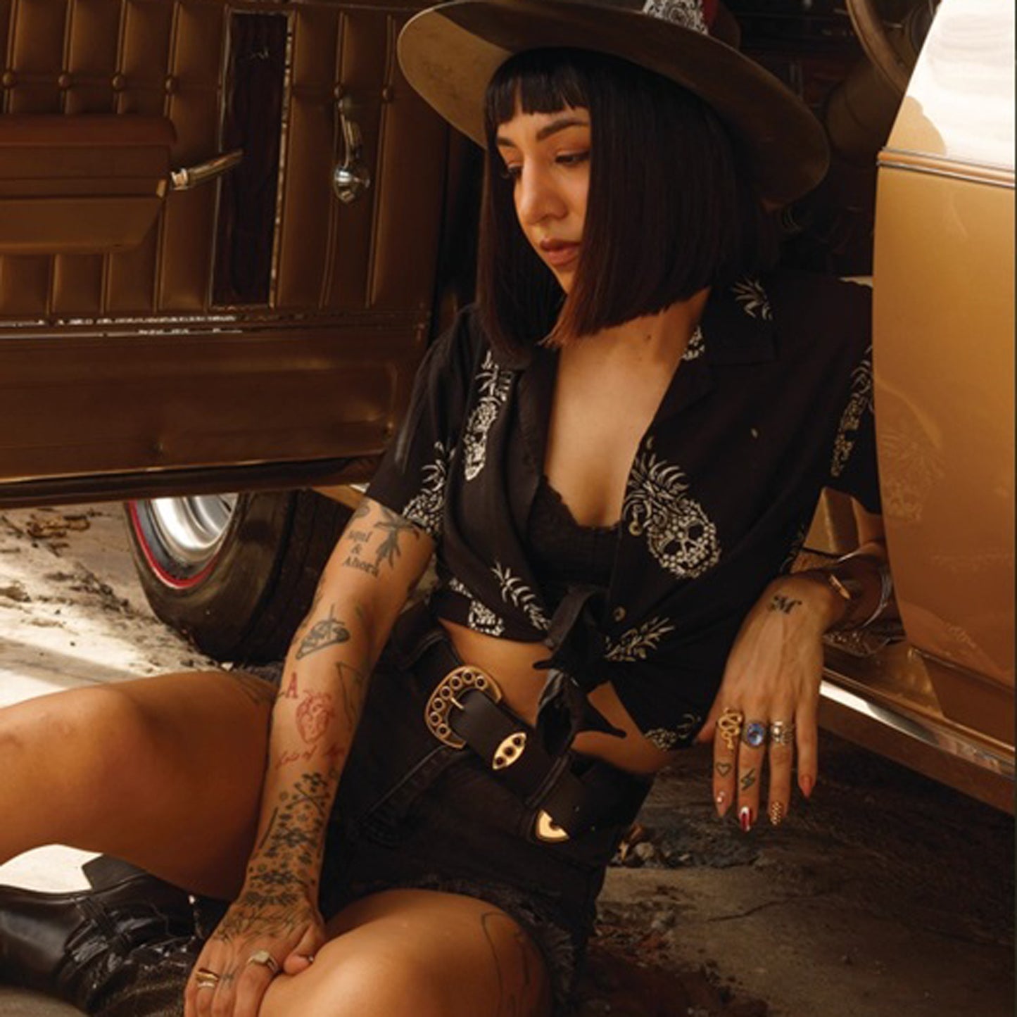 A woman with a black bob haircut and a wide-brim hat sits on the ground next to a vintage car, wearing a black shirt with a white pineapple skull print, black shorts, and a black belt with a silver buckle.