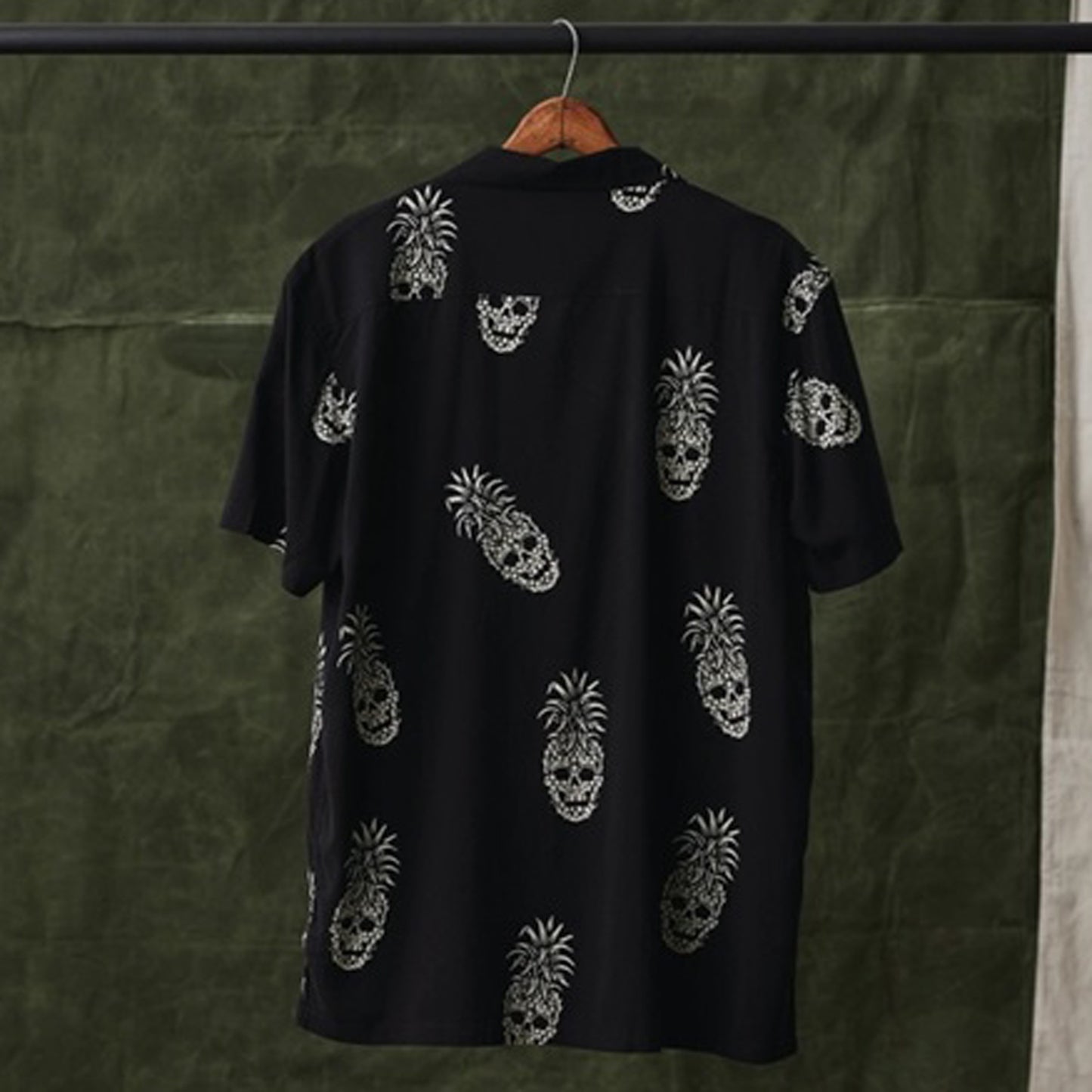 A black short-sleeve button-up shirt hanging on a wooden hanger, featuring an all-over white skull pineapple pattern, set against a dark olive green textured backdrop.