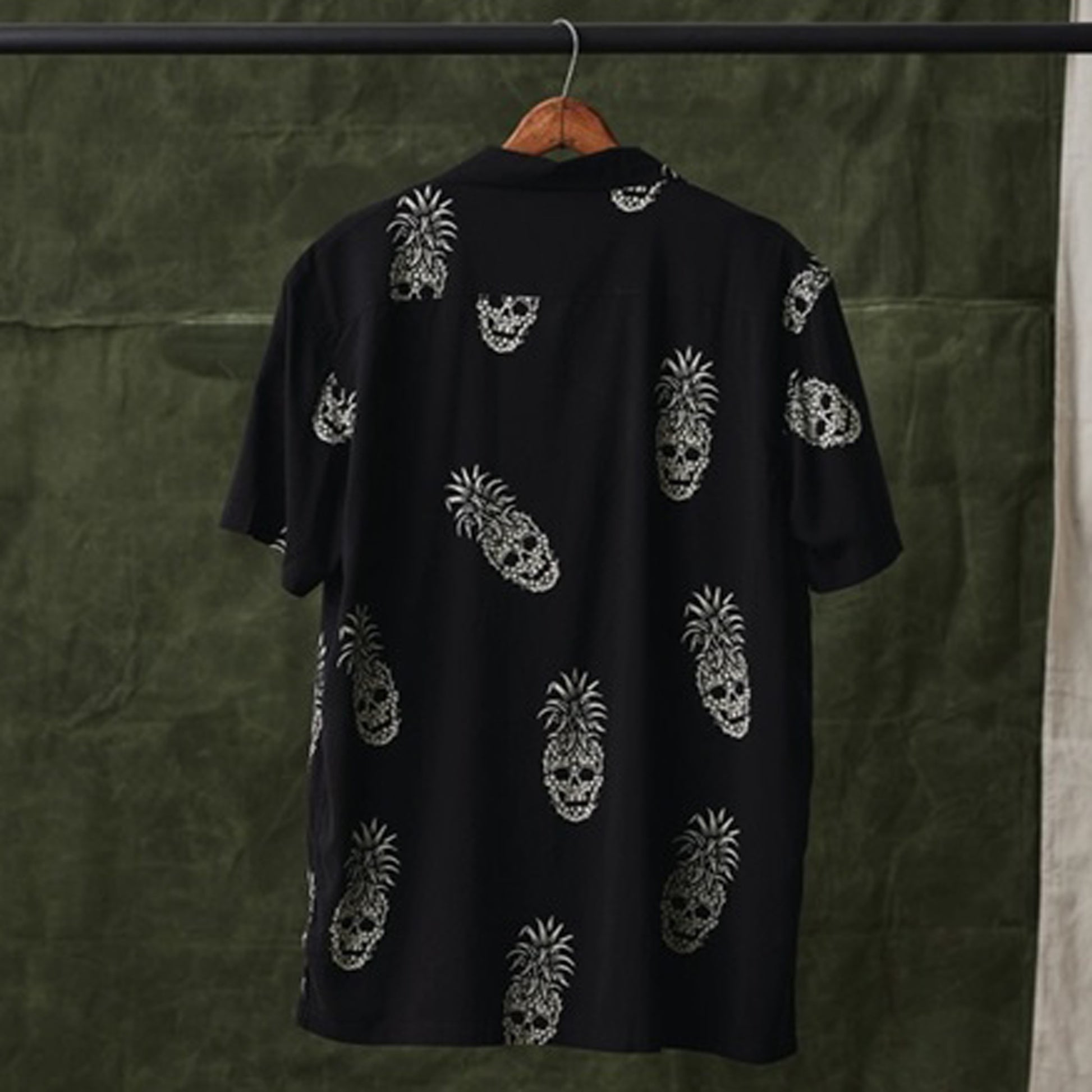A black short-sleeve button-up shirt hanging on a wooden hanger, featuring an all-over white skull pineapple pattern, set against a dark olive green textured backdrop.