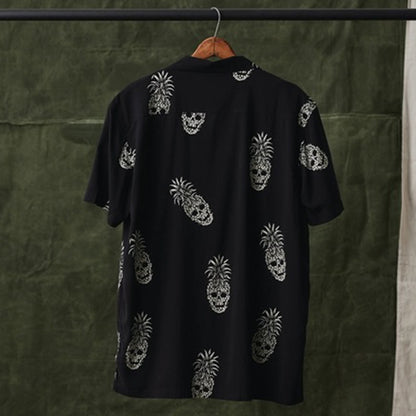 A black short-sleeve button-up shirt hanging on a wooden hanger, featuring an all-over white skull pineapple pattern, set against a dark olive green textured backdrop.
