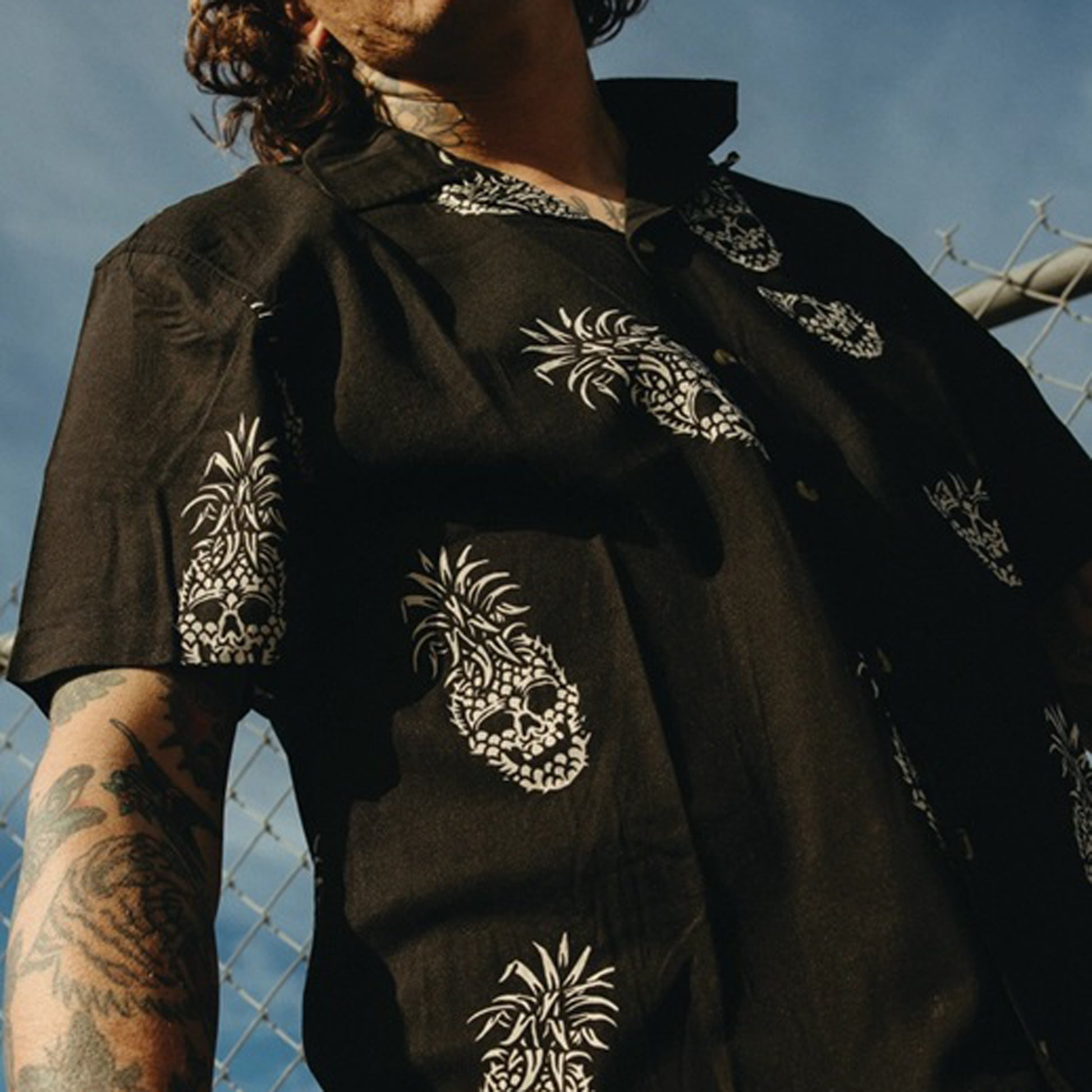 A man wearing a black short-sleeve button-up shirt with white pineapple skull patterns, photographed outdoors against a blue sky and chain-link fence.