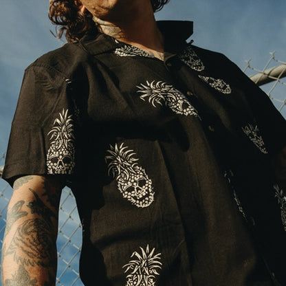 A man wearing a black short-sleeve button-up shirt with white pineapple skull patterns, photographed outdoors against a blue sky and chain-link fence.