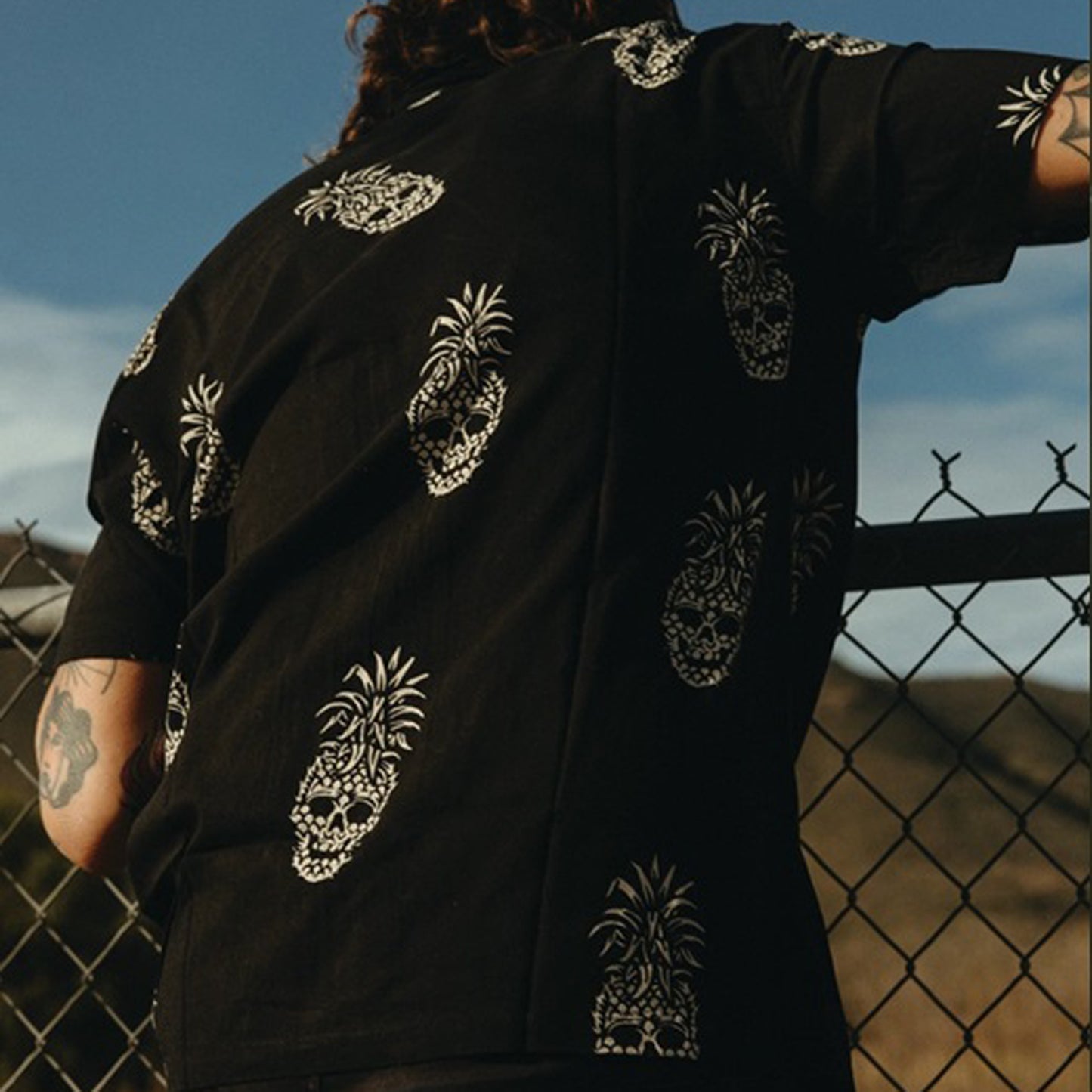 A person wearing a black short-sleeve shirt with white pineapple skull prints, photographed outdoors against a fence and blue sky.