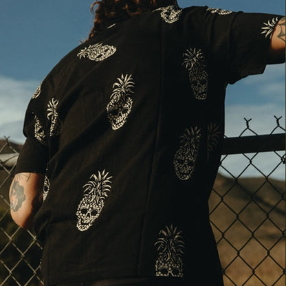 A person wearing a black short-sleeve shirt with white pineapple skull prints, photographed outdoors against a fence and blue sky.