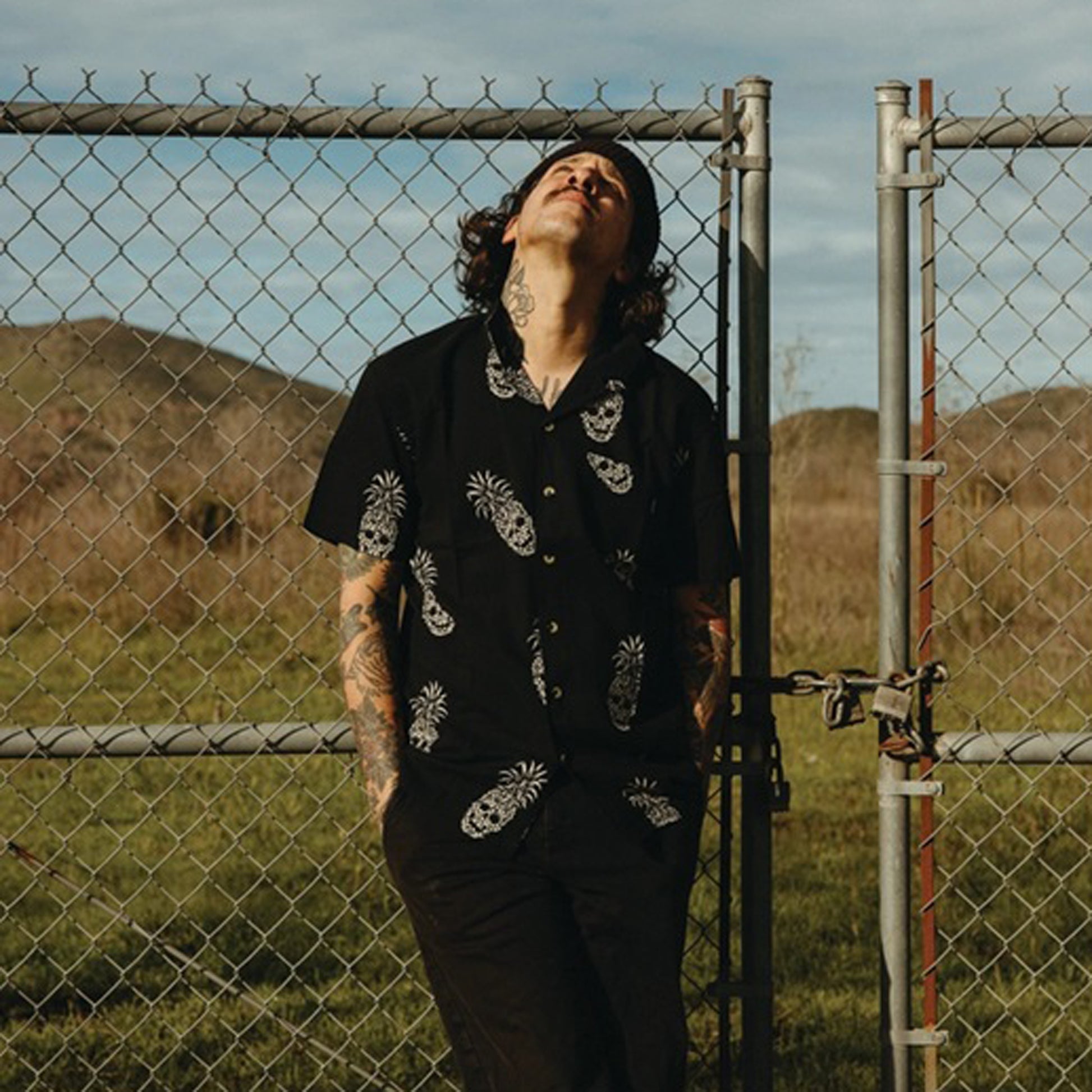 A man with tattoos stands in front of a chain-link fence outdoors, wearing a black short-sleeve button-up shirt with white pineapple skull print design and black pants, looking upward.