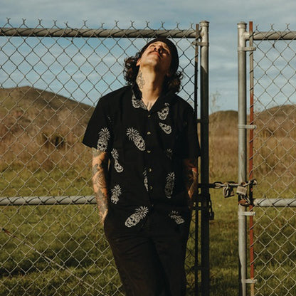 A man with tattoos stands in front of a chain-link fence outdoors, wearing a black short-sleeve button-up shirt with white pineapple skull print design and black pants, looking upward.