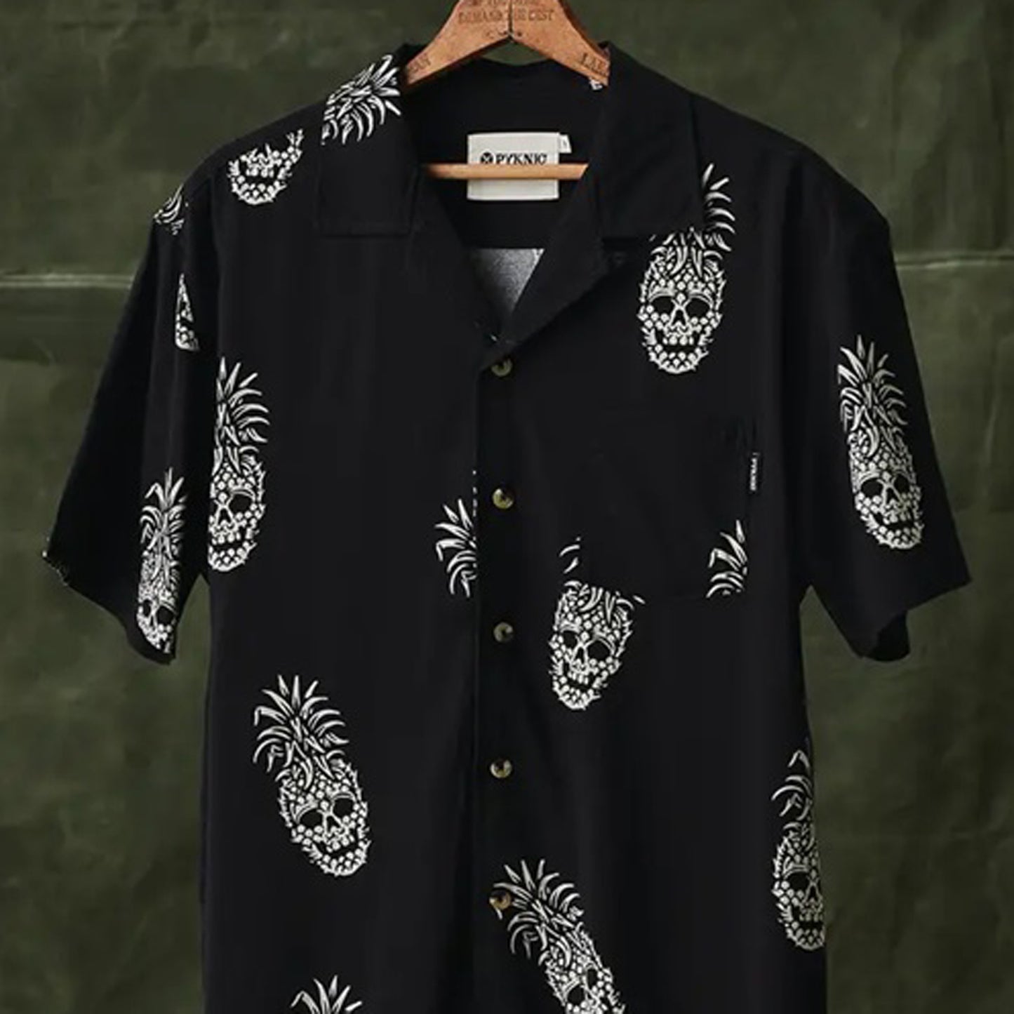 A black short-sleeve button-up shirt with a white skull-pineapple print, hanging on a wooden hanger against a dark olive green background. The brand name 'PYKNIC' is visible on the inner neck label.